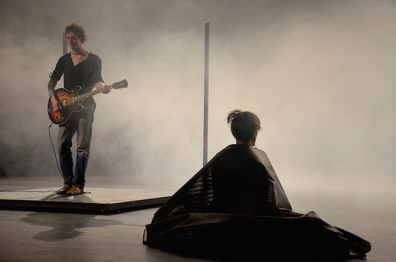 A man stands on a square with a guitar while a performer is enshrouded in a black tarp. Smoke wafts through the air